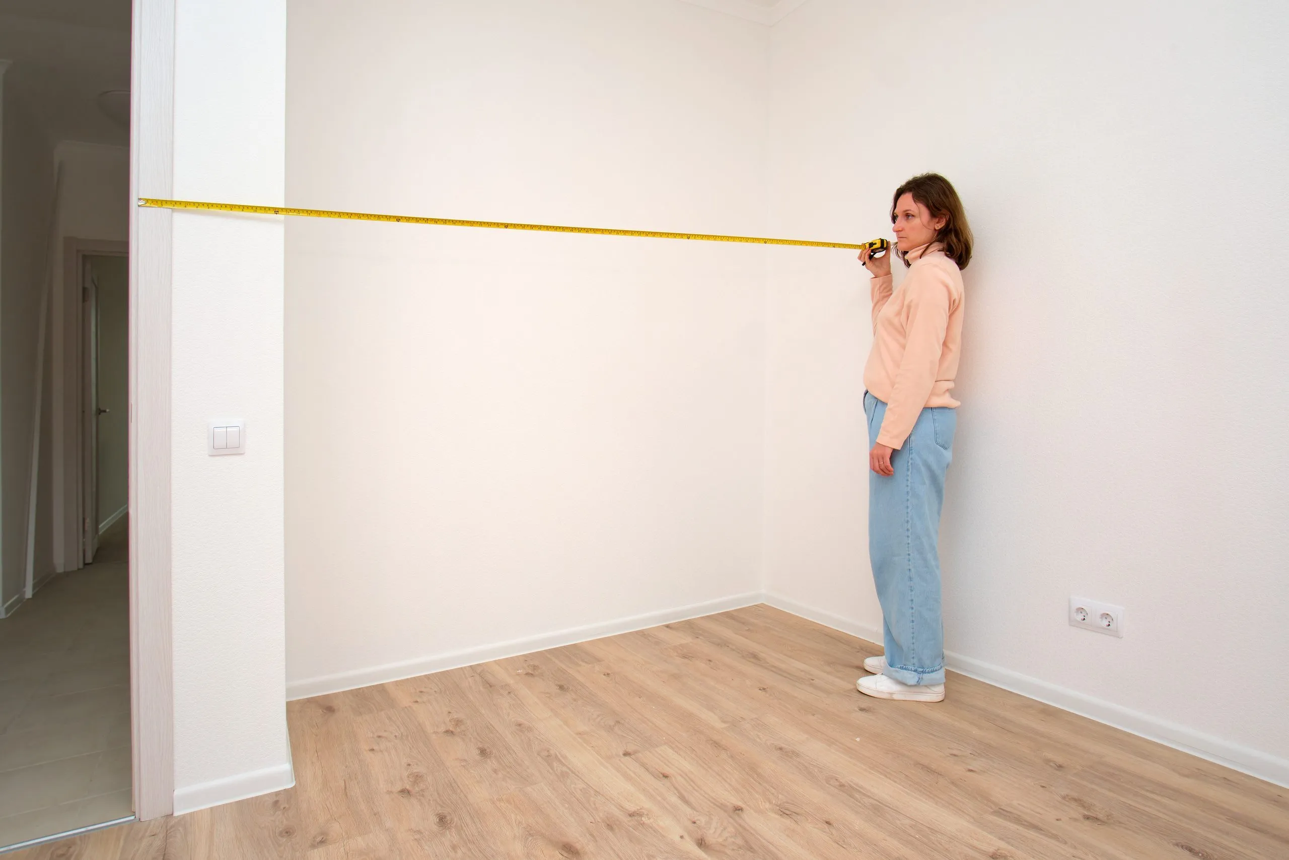 Frau misst mit einem Maßband die Wandlänge in einem leeren Zimmer
